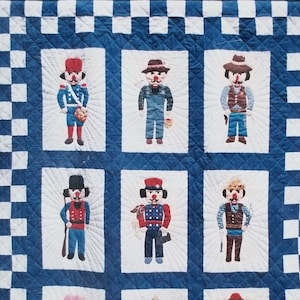 May include: A quilt pattern titled "The Nutcracker Quilt" featuring nine squares with illustrations of nutcracker figures in various outfits. The quilt has a blue and white checkerboard border and a decorative wave pattern.