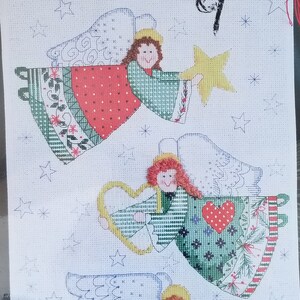 May include: A Christmas Traditions cross-stitch kit featuring a banner with three angels. The angels have red, green, and white outfits, with gold accents. The kit is 10 x 19 inches (25.4 x 48.3 cm) and includes the text "Designs for the Needle".