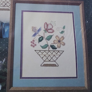 May include: A Counted Bead Embroidery kit featuring a floral design. The kit includes glass beads, cotton floss, Aida fabric, needles, and instructions. The design is of a flower basket, with a framed cross-stitch example and a pillow with the same design.