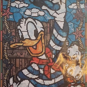 May include: A cross-stitch chart featuring Donald Duck in a stained glass design. The image shows Donald Duck in a blue and white sailor outfit with a red bow tie. The picture size is 10.5" x 15.4". The chart includes a symbol and thread chart.