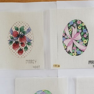 May include: Collection of seven hand-painted Easter egg designs on canvas. Each egg features unique patterns, including strawberries, floral arrangements, and ribbons. The name "Marcy" is printed on some of the canvases. The designs are in various colors.