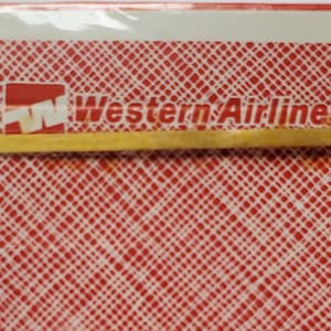 May include: A sealed deck of playing cards with a red crisscross pattern on the back. The words "Western Airlines" are printed in white, along with the airline's logo. A yellow rubber band secures the deck.
