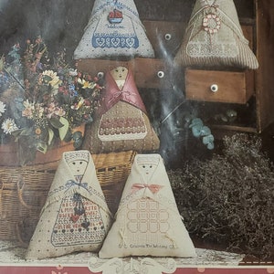 May include: A package for "The Damsels" counted cross stitch and needlepoint designs. The cover is a reddish-brown with a floral pattern. The image shows several doll-like figures with cross-stitched designs, and the text "PRIMROSE COLLECTION" is visible.