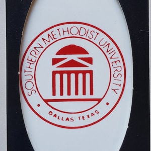 May include: A rectangular box with a white oval-shaped window revealing a white oval-shaped object. The object features the words "SOUTHERN METHODIST UNIVERSITY" in a red circular design, with "DALLAS TEXAS" below. The box has a black background.