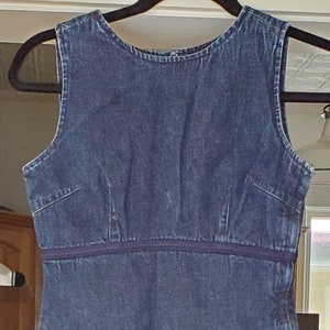 May include: A sleeveless, dark blue denim dress with a fitted bodice and an A-line skirt. The dress features a dark purple band at the waist and a small red tag near the hem. It hangs on a black hanger.