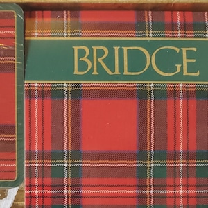 May include: A boxed set of Bridge playing cards. The cards and box feature a red, green, and white plaid pattern. The box has the word "BRIDGE" in gold lettering on a green banner. The set includes two decks of cards and a score pad.
