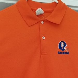 May include: Bright orange polo shirt featuring the Denver Broncos logo on the left chest. The logo includes a blue and orange helmet with the word "BRONCOS" in blue. The shirt has a collared neckline and three white buttons.