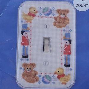May include: A cross-stitch kit for a light switch cover. The design features a border of toy soldiers, teddy bears, and ducks. The words "Toys on Parade" are printed at the bottom. The kit is in a plastic package with the words "Fond Memories" printed on the top.