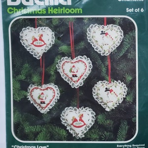 May include: Two Bucilla Christmas Heirloom ornament kits. One kit features heart-shaped ornaments with Christmas themes, the other features shapes and faces. Each kit contains six ornaments. The kits are packaged in clear plastic.
