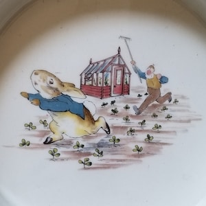 May include: White ceramic plate with a colorful illustration of a rabbit running away from a man in a garden. The plate has text around the rim that reads, "But round the end of a cucumber frame, whom should he meet but Mr. McGregor!"