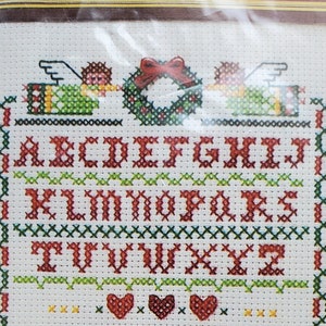 May include: Cross-stitch sampler with a brown frame. The design features the alphabet, the words "GOD BLESS US EVERY ONE", and the numbers "1984". The sampler includes Christmas-themed elements like angels and a wreath.