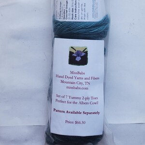May include: A clear plastic package containing seven skeins of hand-dyed 2-ply yarn in a gradient of colors ranging from dark charcoal to teal. Each skein has a white label with text including MissBabs Hand Dyed Yarns and Fibers.