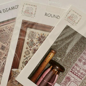 May include: Four cross-stitch pattern packets. The packets are labeled "Quaker Diamond", "Round", and "Claret". The "Claret" packet includes a wooden spool and a small card. The packets are in clear plastic sleeves.