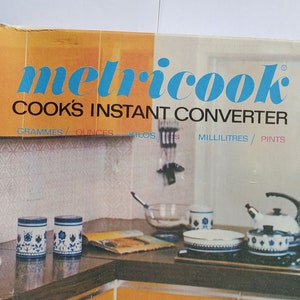 May include: A vintage "metricook" kitchen conversion tool box. The box features the text "Cook's Instant Converter" and includes measurements in grams, ounces, milliliters, and pints. The box also shows a kitchen scene with a stove and kitchenware.