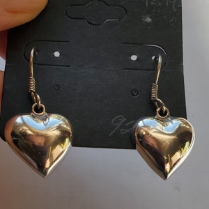 May include: A pair of silver heart-shaped earrings with a polished finish. The earrings are hanging from a black tag. The tag has a price tag and the number 925 written on it.
