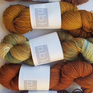 May include: Five skeins of yarn in various colors, including brown, gold, and purple. Each skein has a white label with the letters "nbk" and the words "northbound knitting". The yarn is arranged on a white surface.