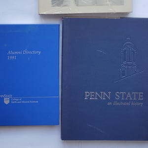 May include: Three books are displayed. The top book is titled "THE COLLEGE OF EARTH AND MINERAL SCIENCES AT PENN STATE" by E. Willard Miller. Below is a blue "Alumni Directory 1991" book. To the right is a navy blue book with "PENN STATE an illustrated history".
