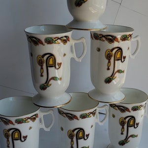 May include: A set of six white ceramic mugs with a pedestal base and a handle. Each mug features a gold rim, a large letter "P" in black and yellow, and colorful Celtic knotwork. The mugs are stacked on top of each other.
