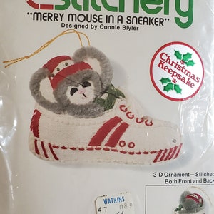 May include: A Jiffy Stitchery kit featuring a "Merry Mouse in a Sneaker" Christmas ornament. The finished size is 3" high. The ornament is a 3-D stitched design of a mouse wearing a red and white sneaker. The kit includes yarn and instructions.