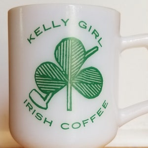 May include: White glass Irish coffee mug with a handle. The mug features the text "KELLY GIRL IRISH COFFEE" in green, along with a green shamrock design. The mug has a pedestal base.