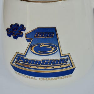 May include: White ceramic mug with a gold rim and handle. The mug features a blue and gold graphic with the text "Penn State Football National Champions 1986". The mug is a standard size for coffee or tea.