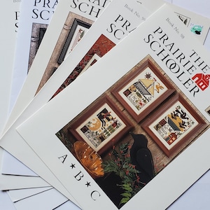 May include: A collection of The Prairie Schooler cross-stitch pattern books. The covers feature framed cross-stitch designs with a variety of images, including a black cat, a butterfly, and a house. The books are fanned out on a white surface.