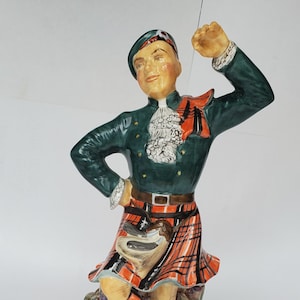 May include: A ceramic figurine of a person wearing a dark green jacket, a red and black plaid kilt, and a matching tam o' shanter hat. The figure stands with one leg crossed, wearing tall socks and black shoes, with a sporran attached to the kilt.
