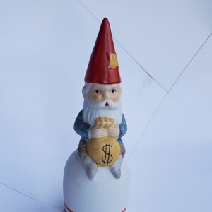 May include: A ceramic gnome figurine with a red hat and blue jacket, holding a money bag with a dollar sign. The gnome sits atop a white base with horizontal stripes. The figurine is approximately 5 inches tall.