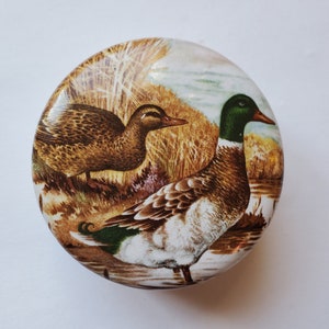 May include: Round decorative tin featuring a detailed illustration of two ducks in a natural wetland setting. The ducks are depicted with brown, green, and white plumage, set against a backdrop of reeds and water. The tin has a glossy finish.