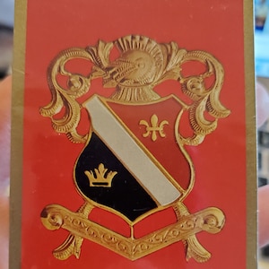 May include: A red playing card deck featuring a gold-colored crest with a knight's helmet, shield, and decorative flourishes. The crest includes a crown and the text "FRED PETERS WELDING SUPPLY CO."