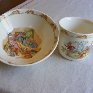 May include: A ceramic bowl and mug set featuring illustrations of rabbits in a whimsical scene. The bowl has a border of running rabbits, and the mug depicts rabbits in a forest setting. The set is likely for children.