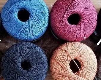 Op de afbeelding: Acht bollen garen in verschillende kleuren, waaronder blauw, rood en oranje, zijn gerangschikt in een doorzichtige plastic zak. Het garen lijkt een fijn gewicht te hebben, geschikt voor brei- of haakprojecten. Het garen heeft een lichte glans.
