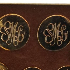 May include: A set of gold-toned buttons with an ornate monogram design, arranged in a brown velvet-lined box. The buttons vary in size and shape, with a shiny, reflective surface.
