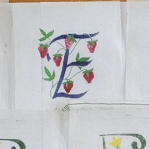 May include: White fabric squares with embroidered letters. The letters are decorated with floral and fruit designs. The letter "A" is adorned with orange and yellow flowers. The letter "E" is decorated with strawberries.