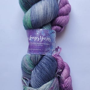 May include: A skein of hand-dyed yarn in shades of blue, green, and purple. The yarn is wrapped with a label that reads "Loops Yarns" and "Dyed yarn created in the Midwest". A smaller skein of purple yarn is next to the larger skein.