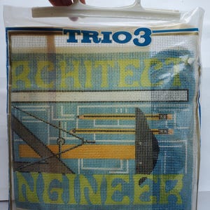 May include: A needlepoint canvas kit in a clear plastic bag with a white handle. The canvas features a blue architectural blueprint design with yellow pencils, a black drafting tool, and the text TRIO 3 ARCHITECT ENGINEER printed in yellow.