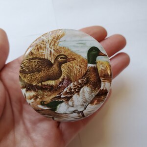 May include: A round, decorative object featuring a detailed illustration of two ducks in a natural setting. The ducks are depicted with brown, green, and white plumage, set against a backdrop of reeds and water. The object has a glossy finish.