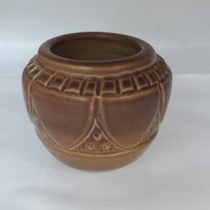 May include: A round, brown ceramic pot featuring a smooth, glazed finish. The exterior is decorated with a repeating embossed pattern of vertical rectangular segments around the rim and a series of rounded, petal-like shapes below.