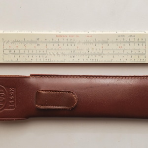 May include: A vintage slide rule with a cream-colored body and black and red markings, labeled "Frederick Post Co." and "Hemmi Japan." It rests above a brown leather case embossed with "POST" and "1444K."