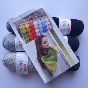 May include: A collection of Katia Ombre yarn skeins in various shades of gray and black, with a brochure featuring a woman wearing a green and yellow scarf. The brochure also displays a color palette of yarn samples.