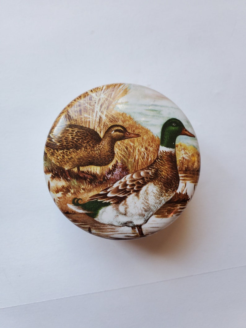 May include: Round decorative tin featuring a detailed illustration of two ducks in a natural wetland setting. The ducks are depicted with brown, green, and white plumage, set against a backdrop of reeds and water. The tin has a glossy finish.