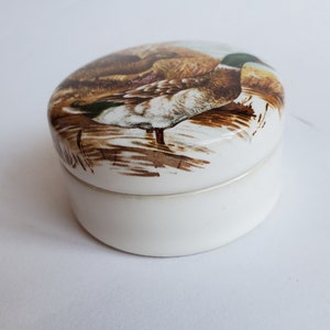 May include: A white, oval-shaped trinket box with a lid featuring a detailed illustration of a duck in a naturalistic setting. The duck has brown, white, and green plumage. The box is made of ceramic or porcelain.