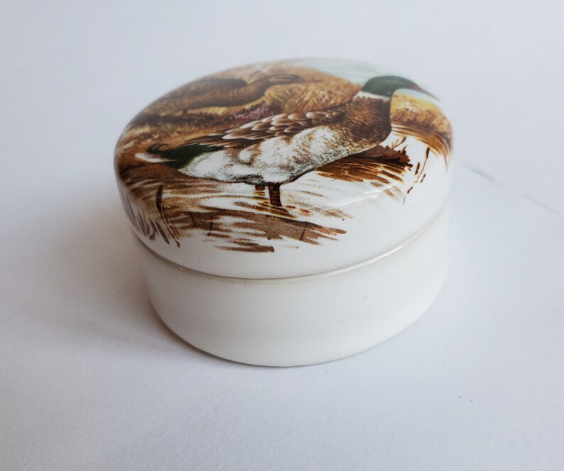 May include: A white, oval-shaped trinket box with a lid featuring a detailed illustration of a duck in a naturalistic setting. The duck has brown, white, and green plumage. The box is made of ceramic or porcelain.