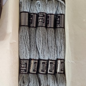 May include: A box of gray D-M-C tapestry wool skeins. Each skein is labeled with the text "Laine tapisserie D-M-C" and "8 metres". The skeins are neatly arranged inside a cardboard box, ready for crafting projects.