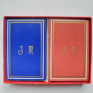 May include: A set of playing cards in a red box. One deck is blue with gold trim and the letters "JM", the other is red with gold trim and the letters "JM". The box is red.