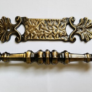 May include: Two antique bronze cabinet hardware pieces. One is a rectangular backplate with ornate floral accents and a textured surface. The other is a matching pull handle with a detailed, symmetrical design.