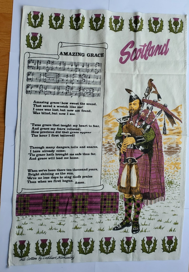 May include: Cream-colored cotton tea towel featuring the text "Amazing Grace" and a Scottish bagpiper. The towel has a purple and green thistle design along the top and bottom edges, and the word "Scotland" in pink.