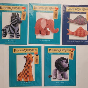 May include: Five fabric animal pattern packets. The packets feature a zebra, elephant, fish, giraffe, and lion. Each packet has a blue border with the text "RUMPLED QUILT SKINS" and the animal's name.