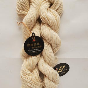 May include: Two skeins of off-white yarn with tags. The tags are black with gold text and a red and gold emblem. The text on the tag reads "EISAKU NORO, the world of nature, Made in Japan" in English and Japanese.