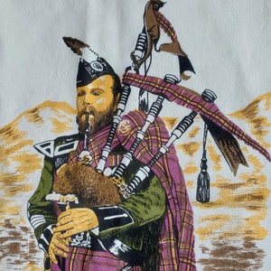May include: A printed image of a Scottish bagpiper in traditional attire. The bagpiper is wearing a plaid kilt and cape in shades of purple, green, and yellow. The background features a landscape with yellow and brown tones.